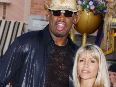 Dennis Rodman is wearing a cowboy hat, shades, and a black leather jacket as Annie Bakes is standing with him.